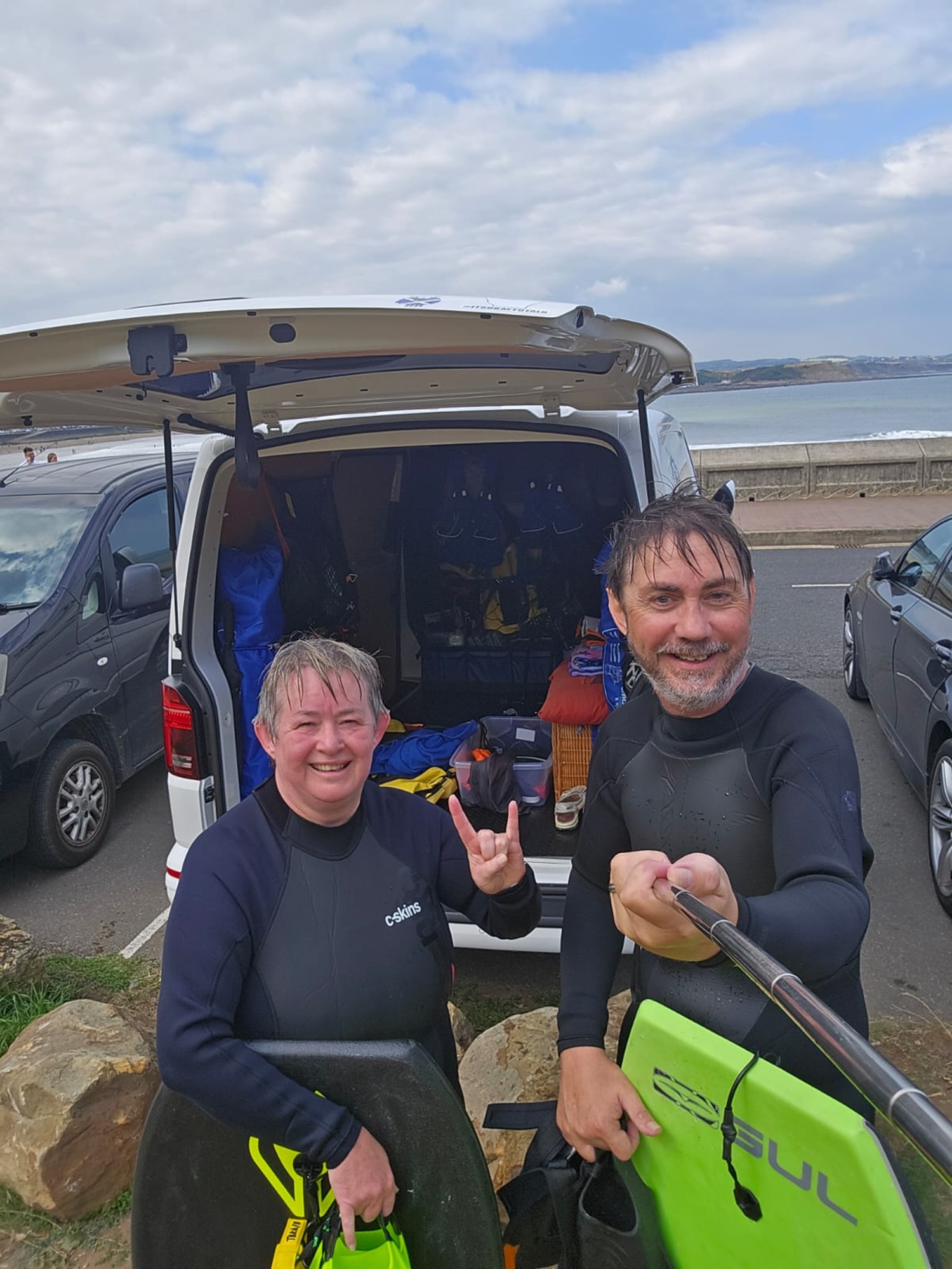 Two surfers in wetsuits by a van at Whitby beach, smiling