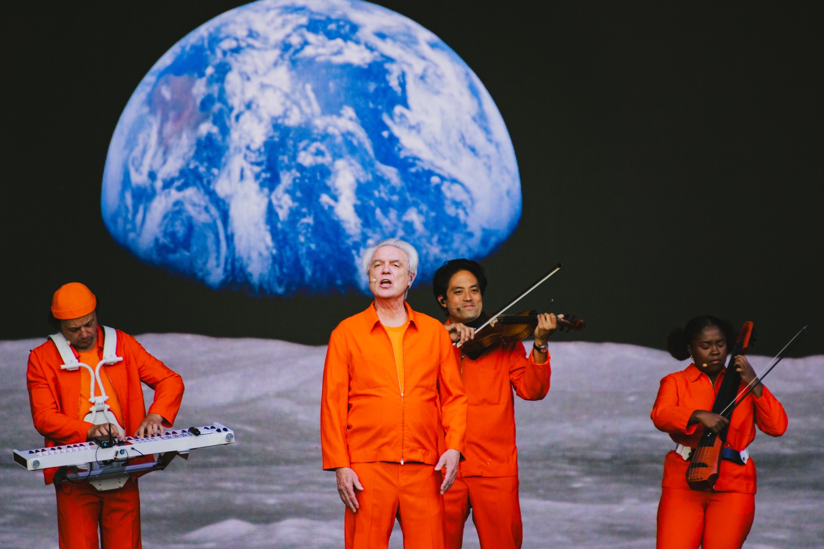 Performers in orange attire on stage with Earth backdrop