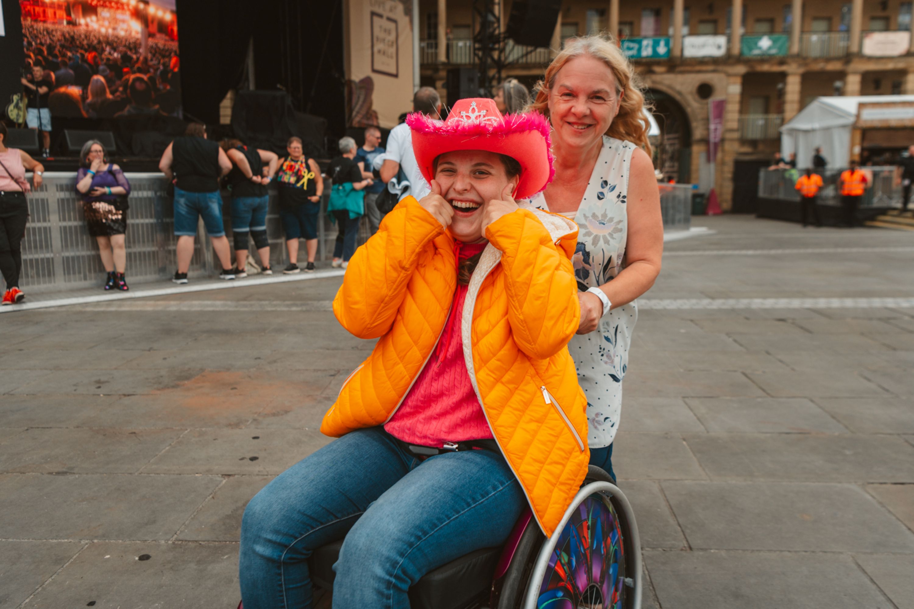Smiling woman in pink cowboy hat and orange jacket in wheelchair at outdoor concert