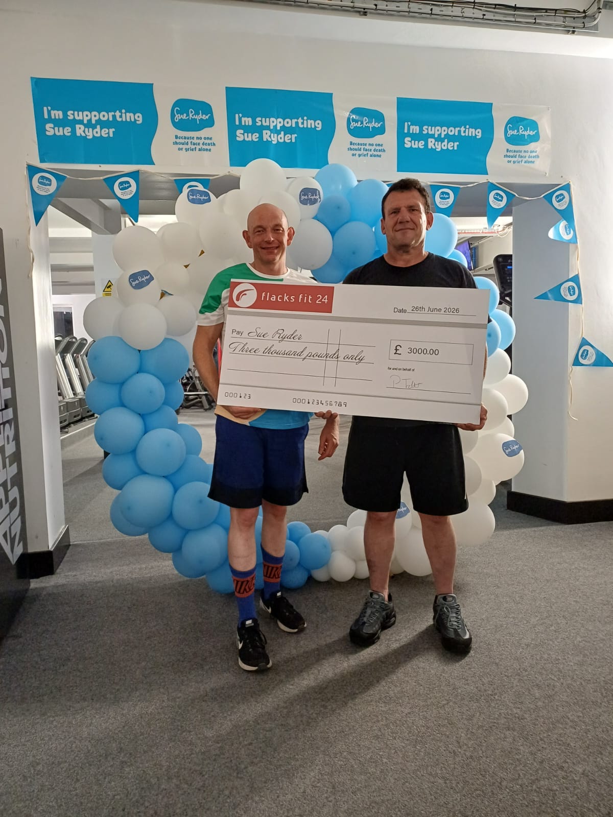 Two men holding a large check in front of blue and white balloon arch