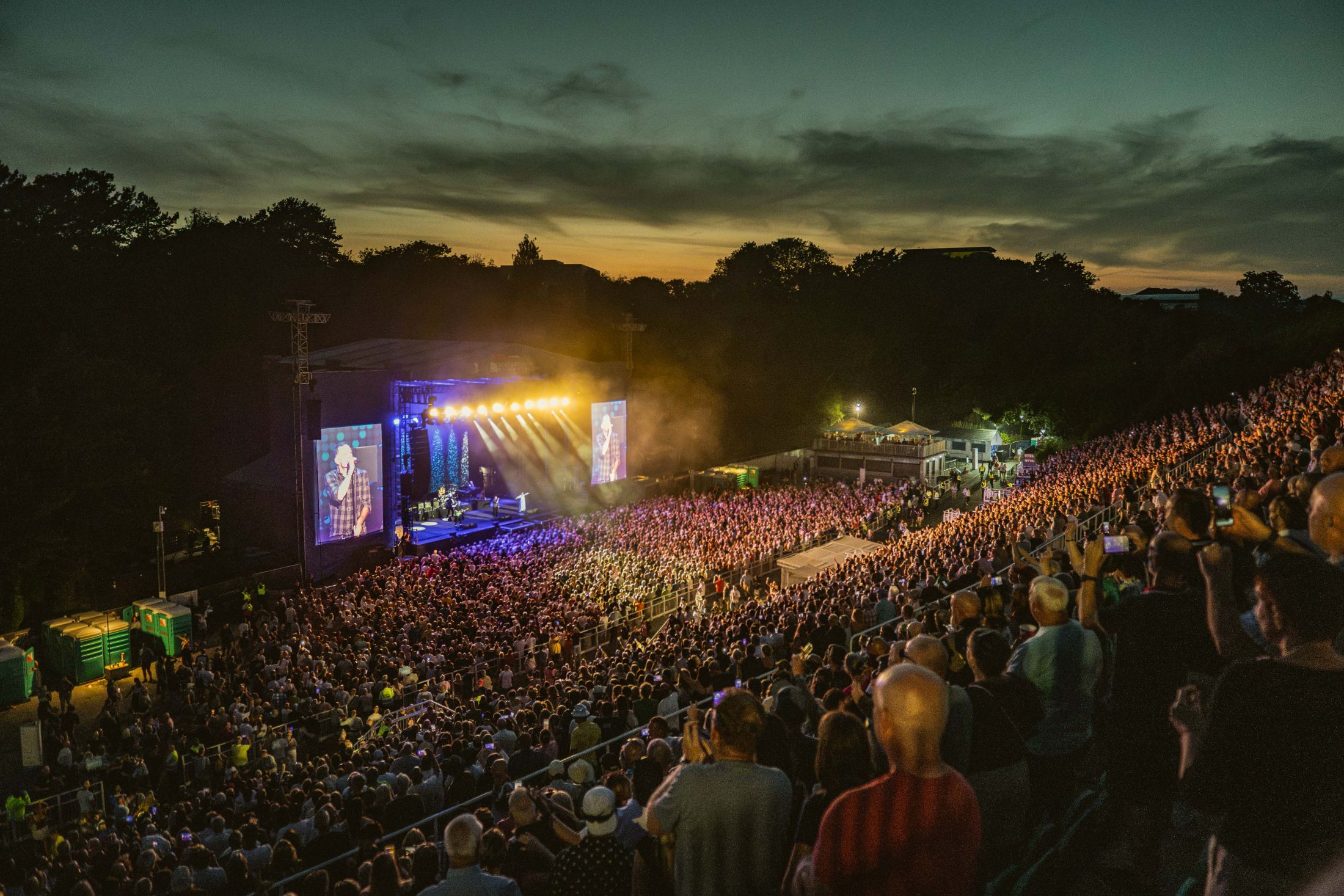 Open-air concert at dusk with large crowd and glowing stage