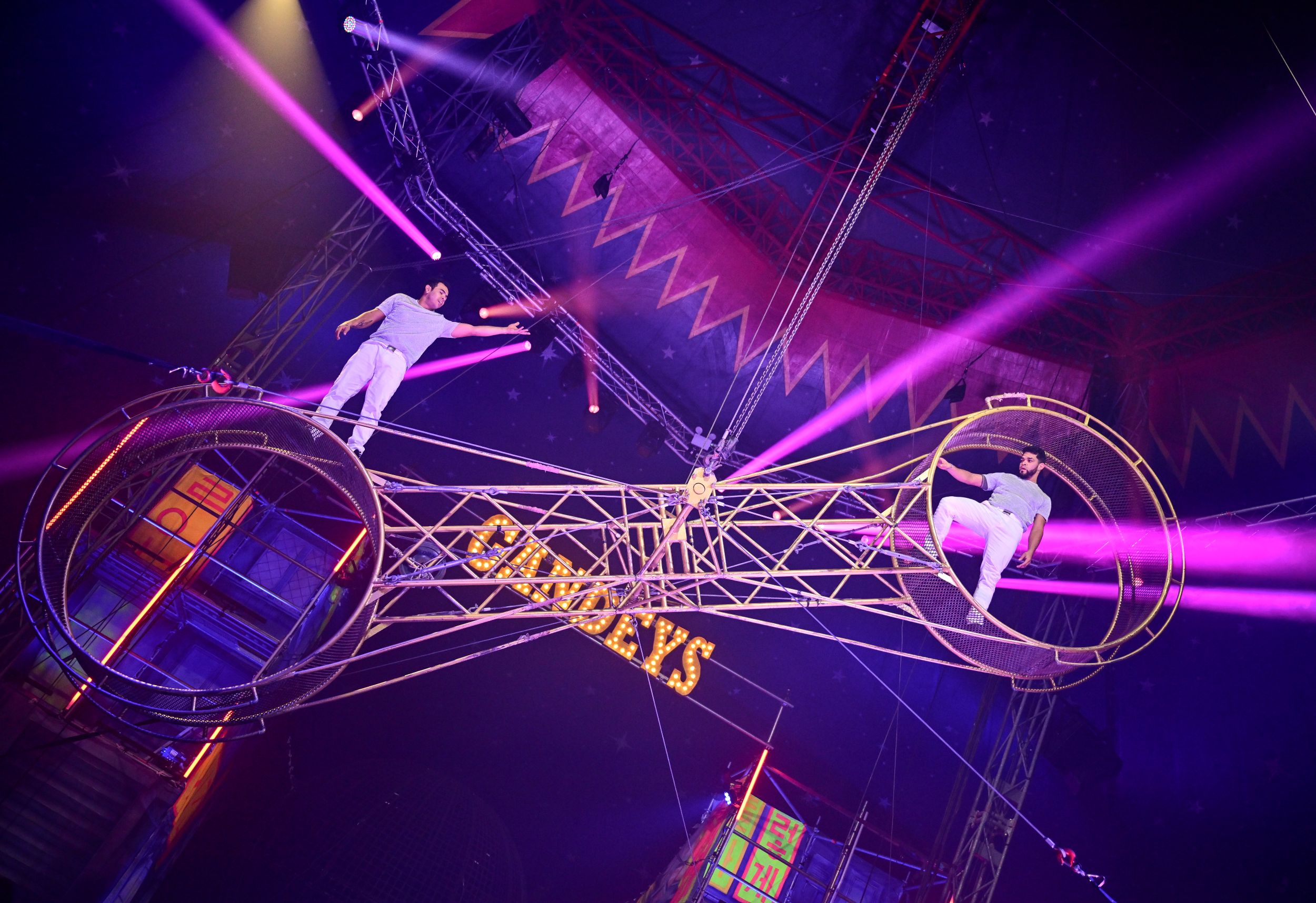 Two acrobats performing on a rotating wheel under pink lights