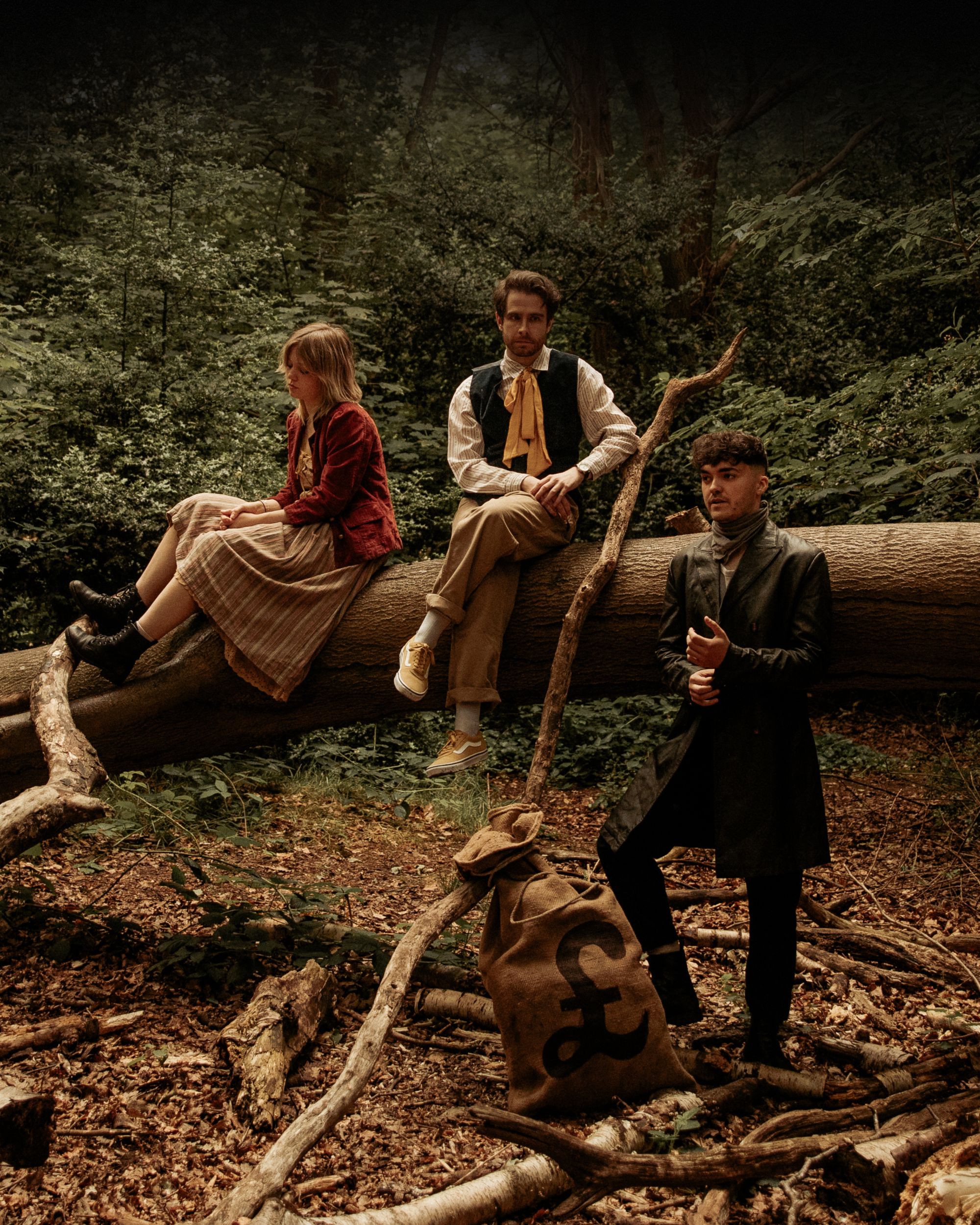 Three people in period costumes on a fallen tree in a forest