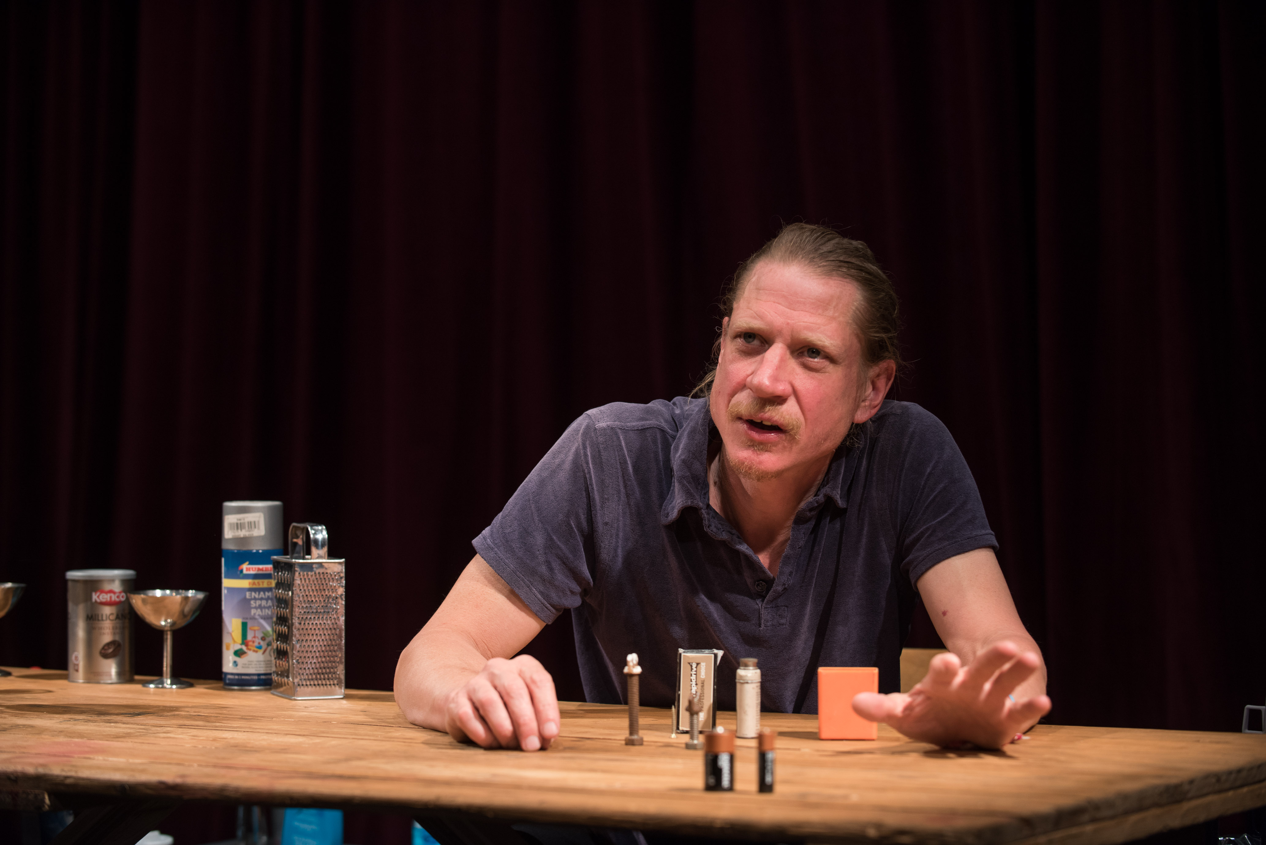 Man speaking at a table with small objects and a dark curtain backdrop