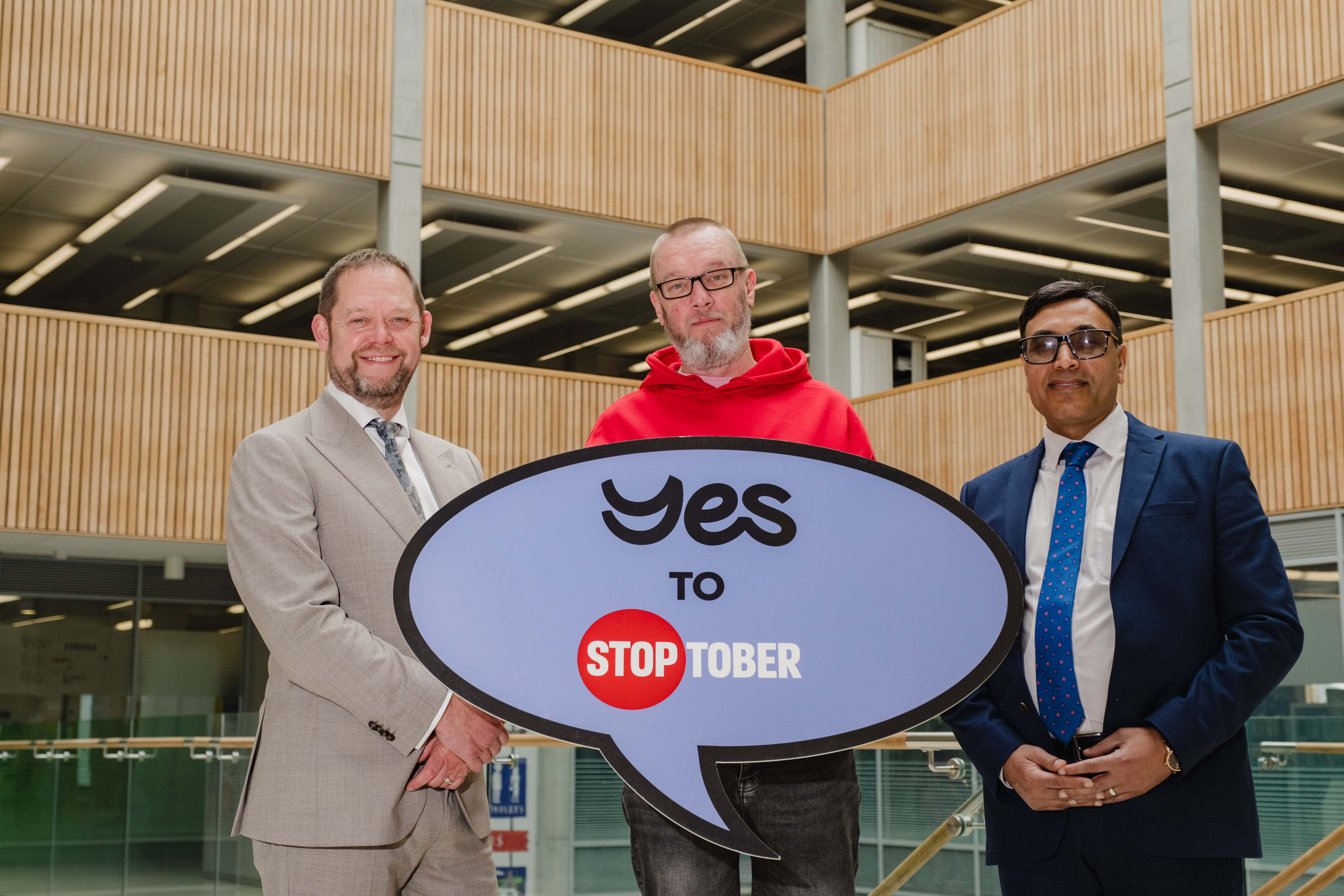 Three men standing indoors holding a 'Yes to Stoptober' sign