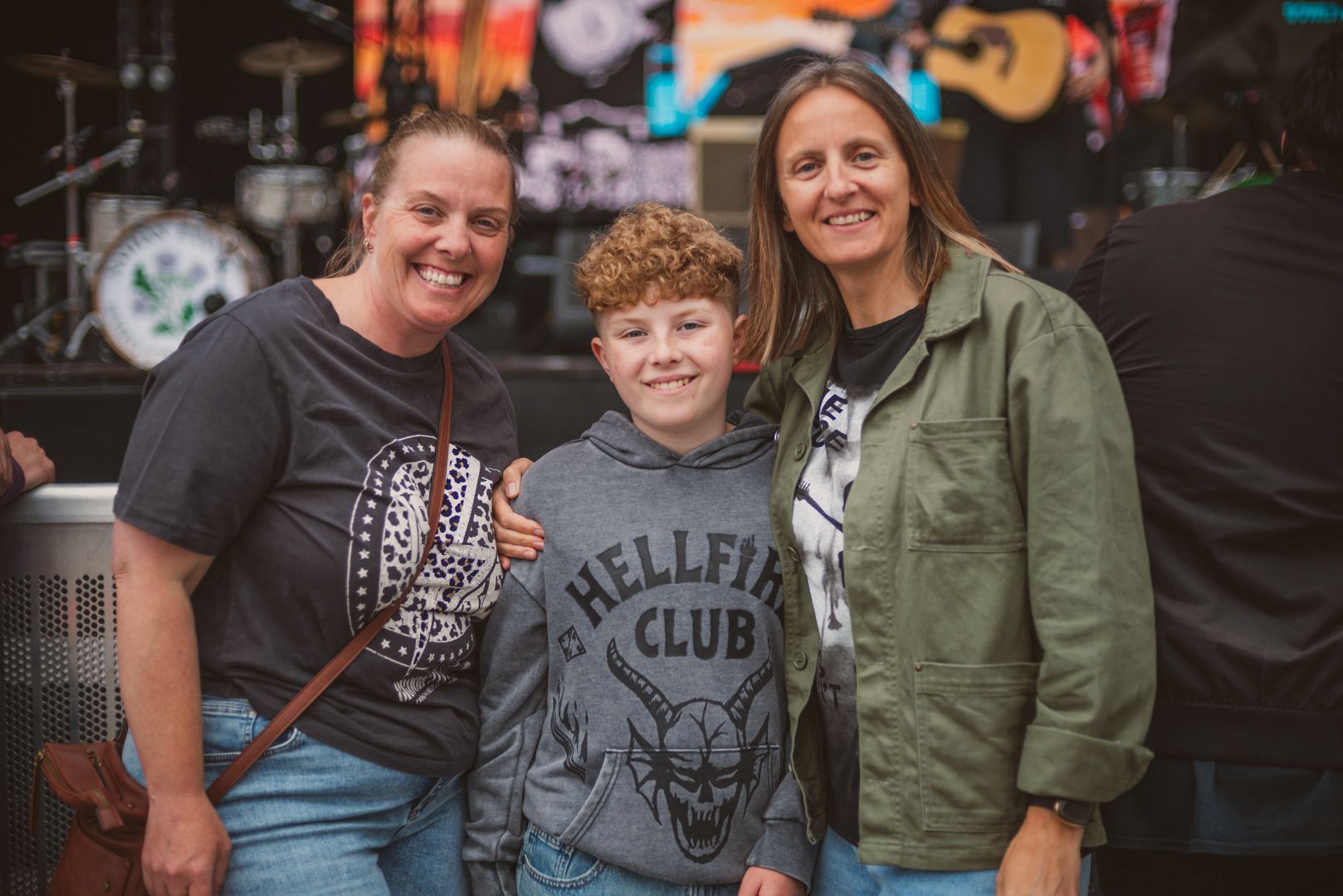 Three people smiling at an outdoor music event with stage behind