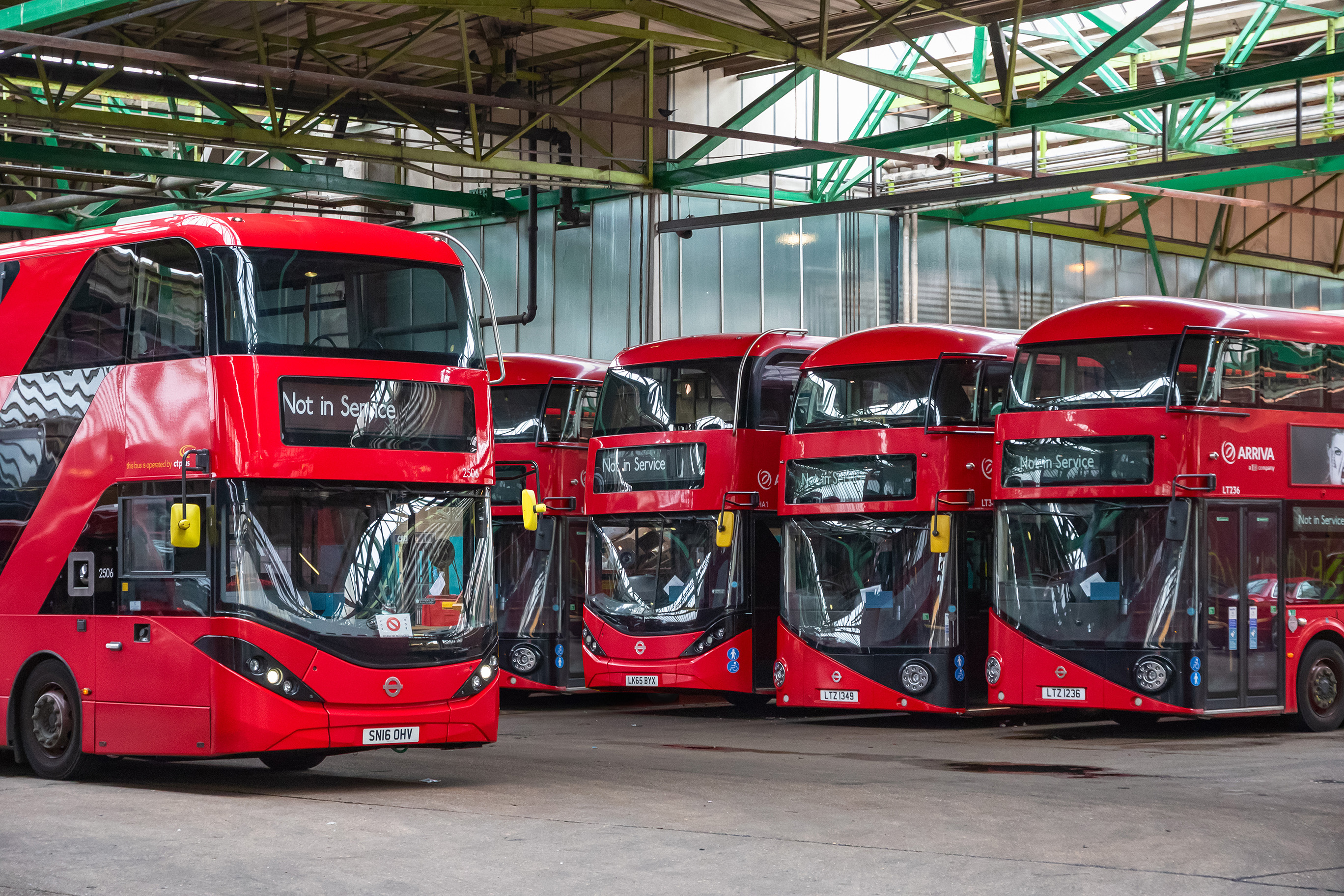 Several red double-decker buses are parked inside a large bus depot with a high metal roof and exposed steel beams. 