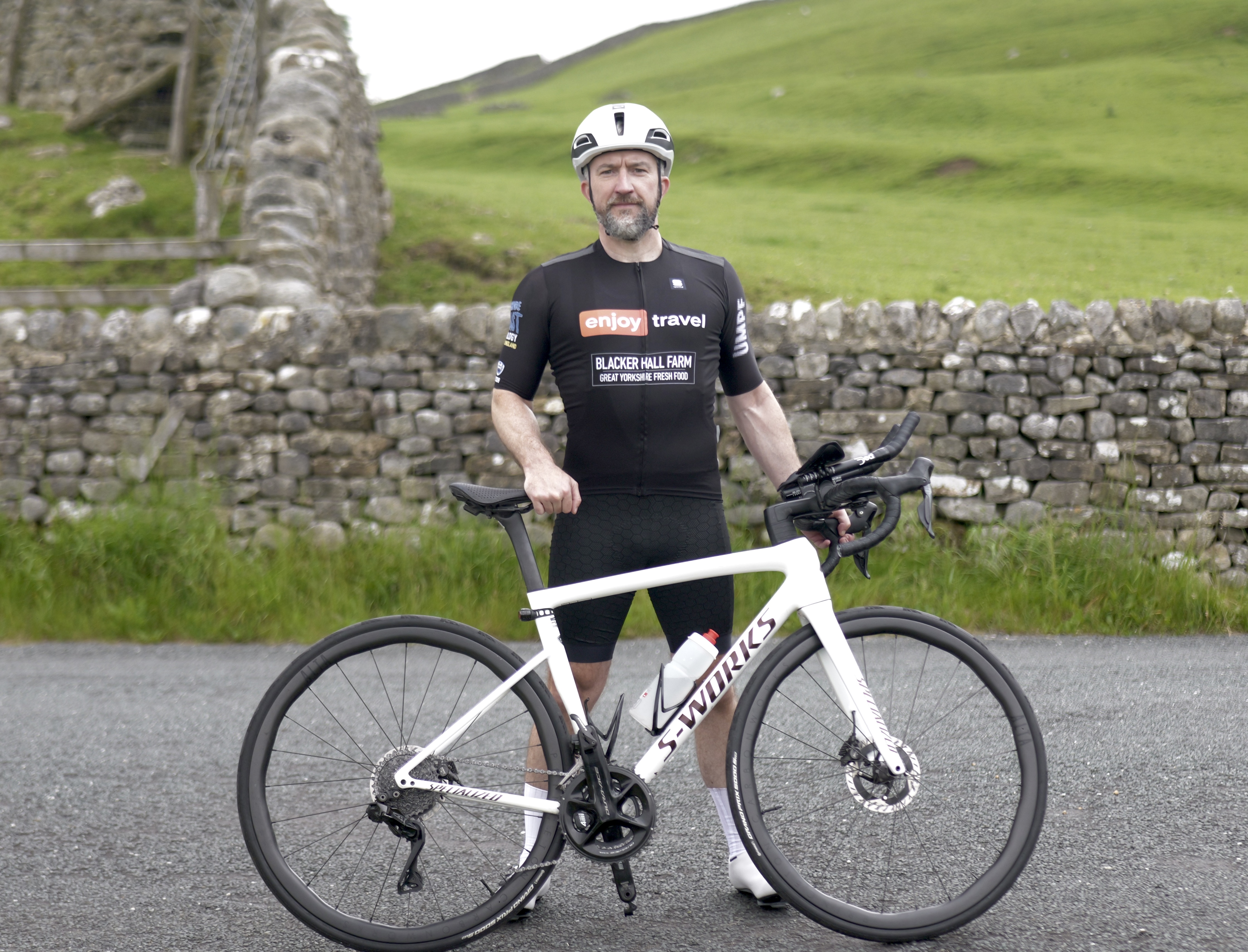 Cyclist in black gear stands with bike near stone wall