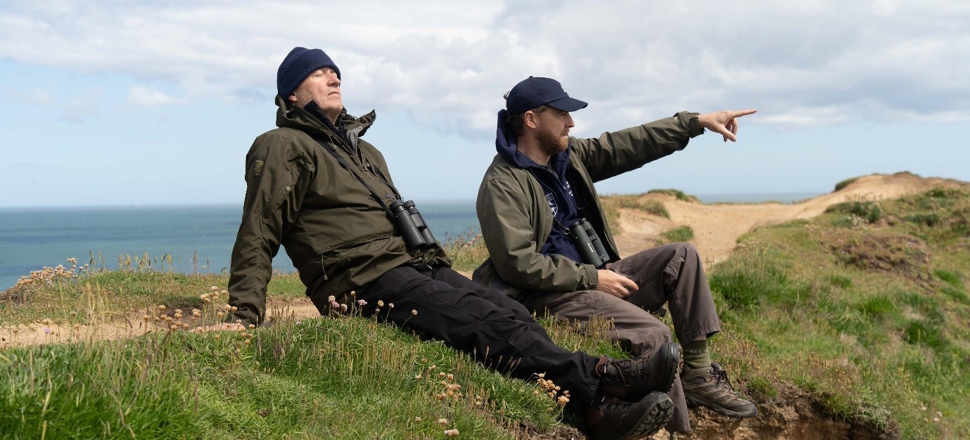 Two men with binoculars on coastal cliff path, pointing at sea