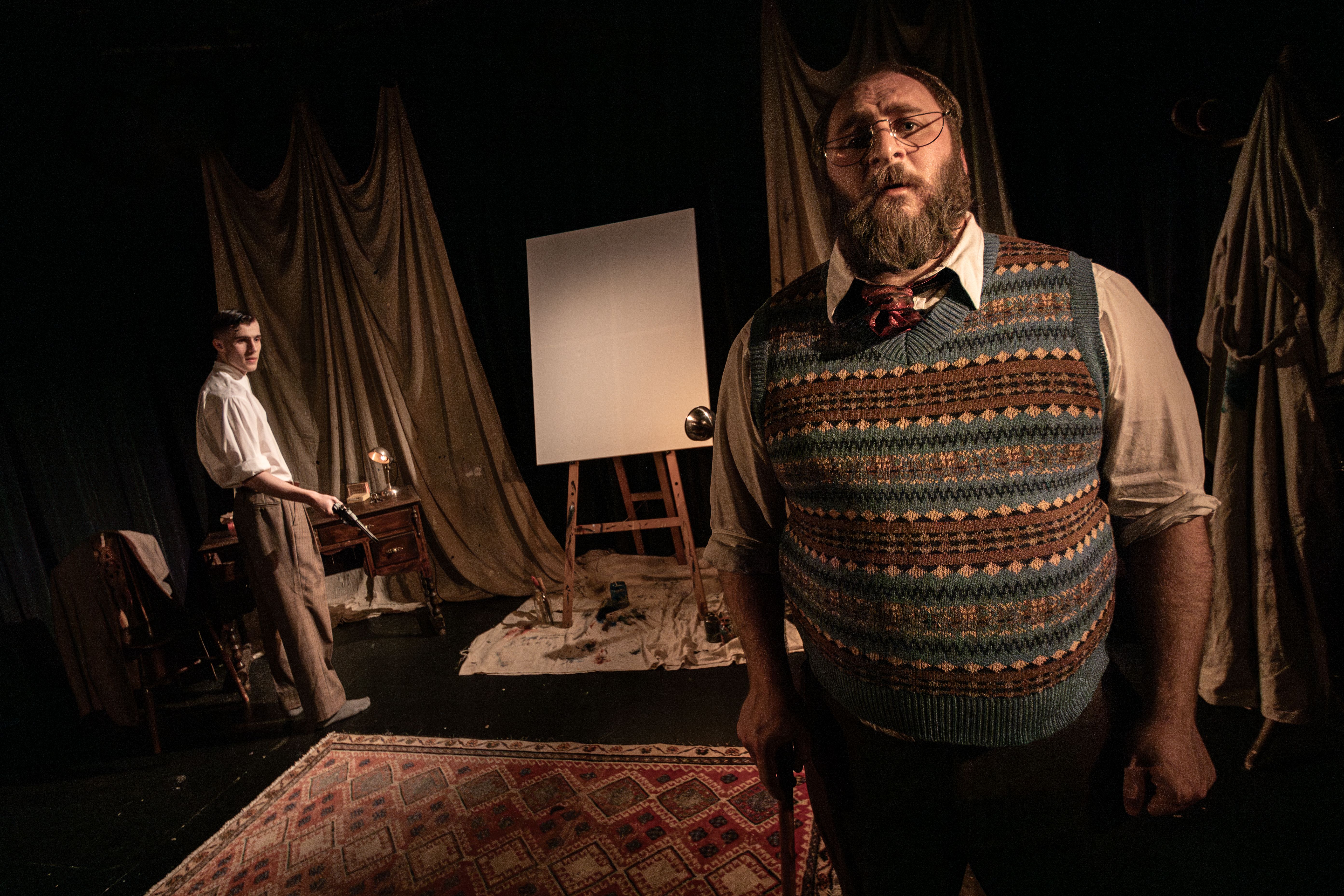 Man in patterned jumper stands in dimly lit room with easel and draped curtains