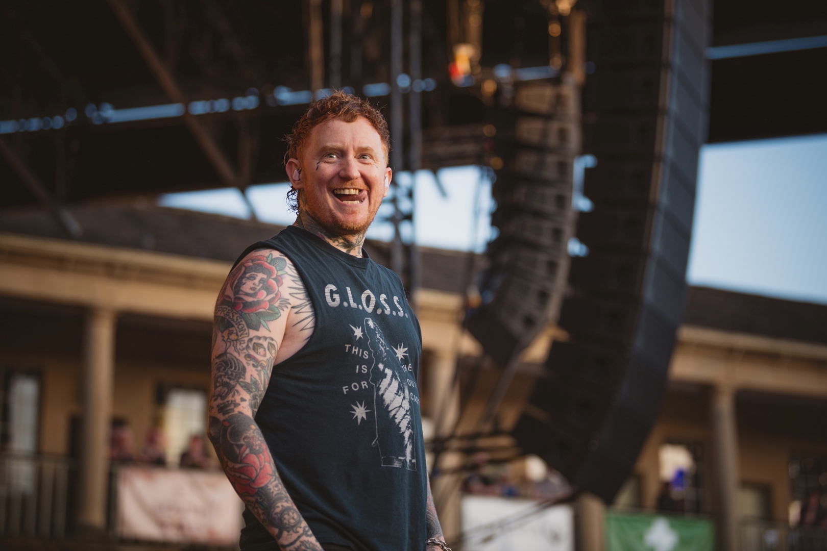 Tattooed musician smiling on outdoor stage with speakers