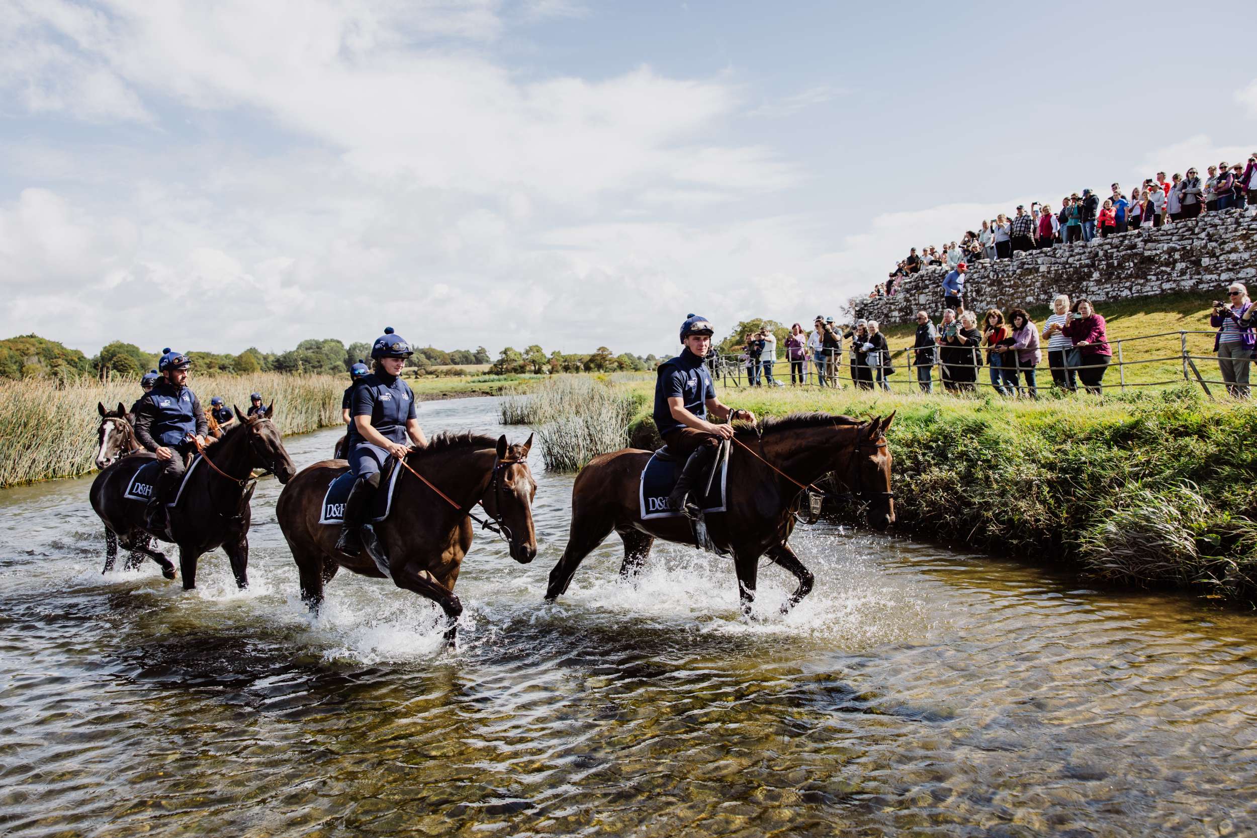 Three riders on horseback crossing a shallow river with spectators on the bank