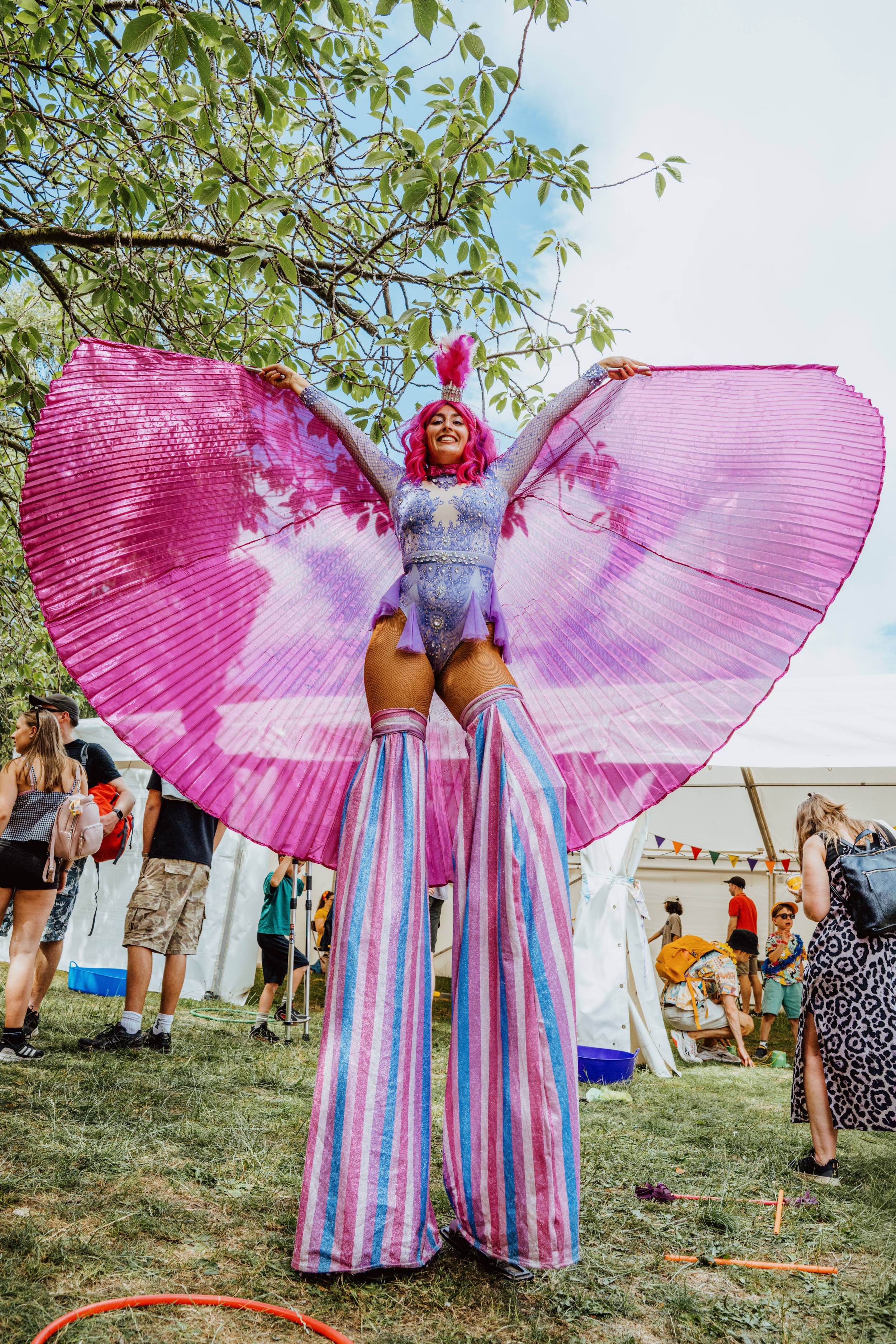 Festival performer on stilts with vibrant pink wings in park