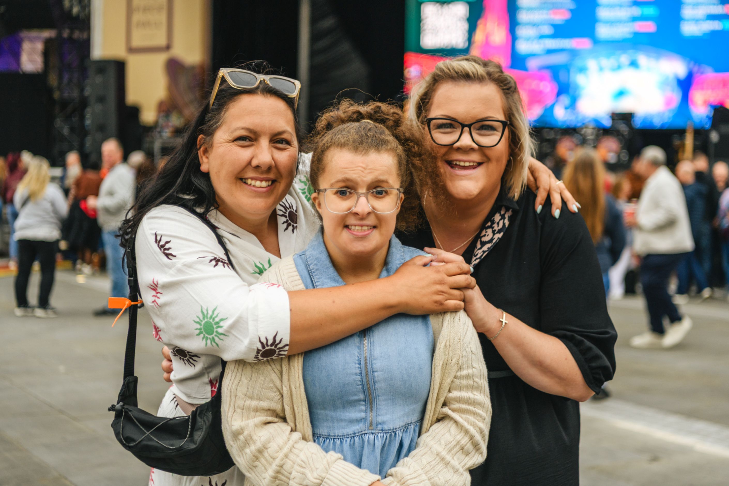Three women smiling at an outdoor festival with a stage in the background