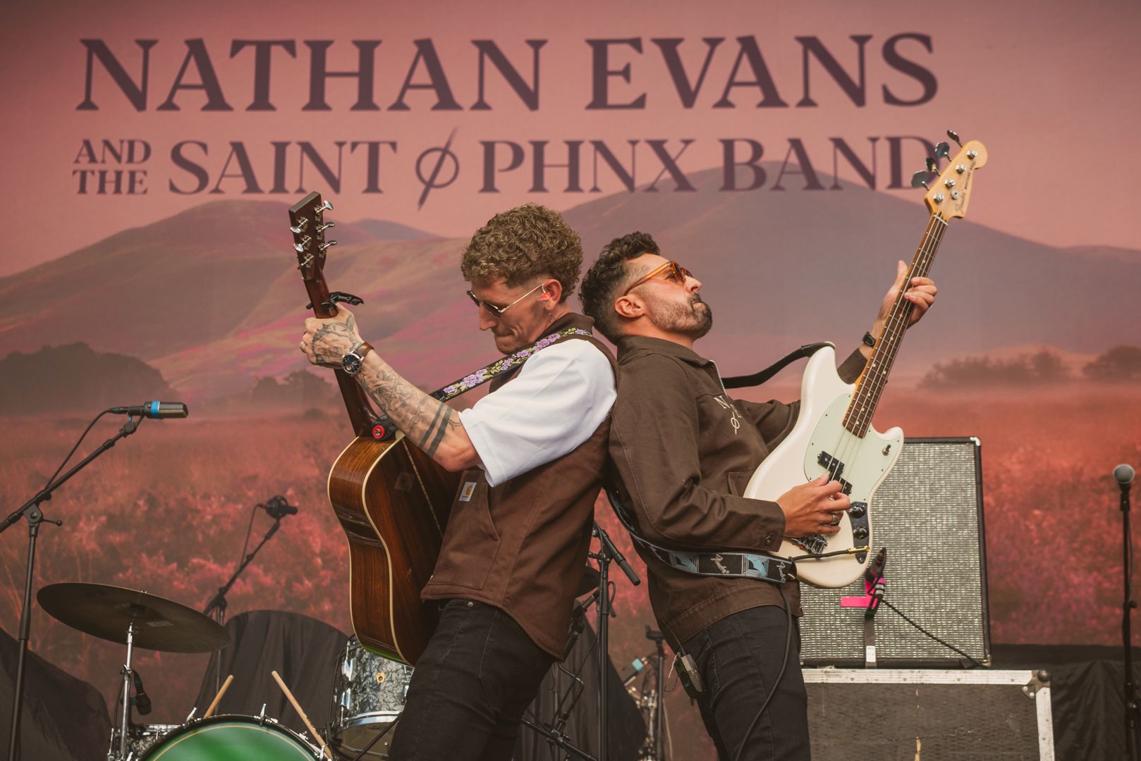 Two musicians performing on stage with guitars against scenic backdrop