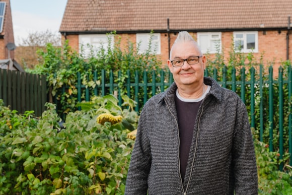 Smiling person in grey coat standing in lush garden with brick houses