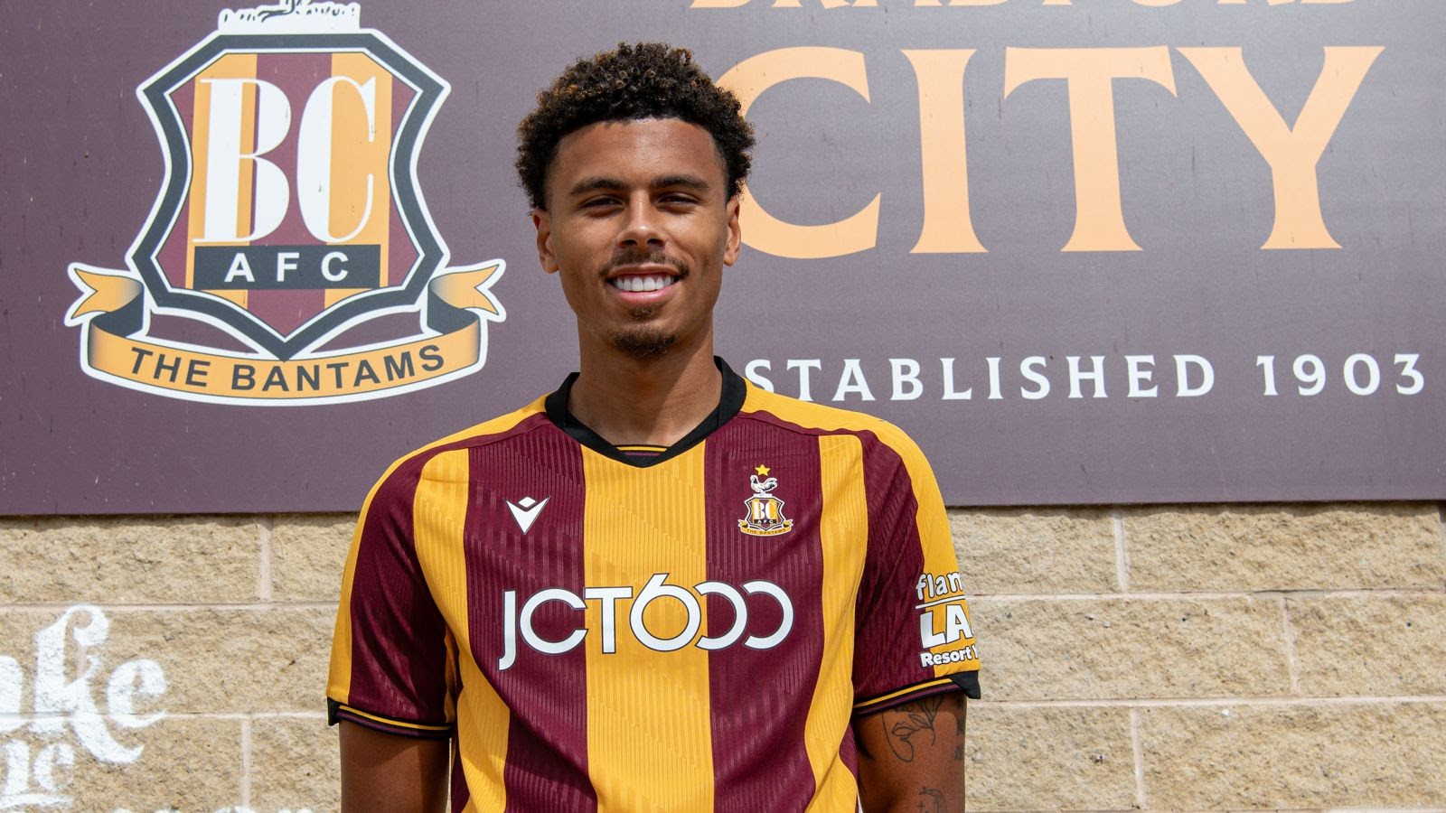 Bradford City AFC player in claret and amber kit standing in front of club sign