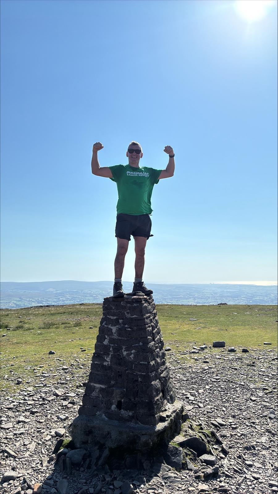 Man in green shirt triumphantly standing on stone cairn on hilltop