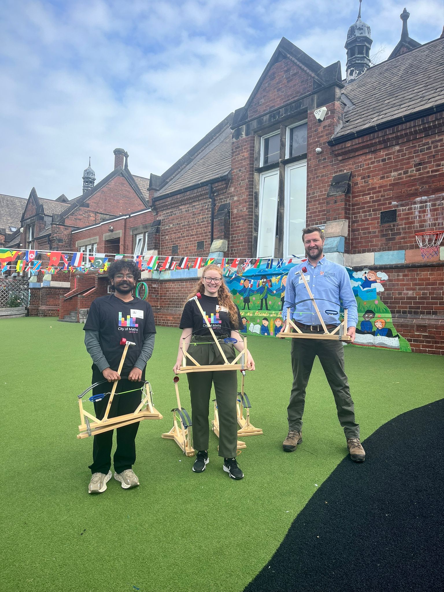 Three people holding wooden catapults in front of a brick building