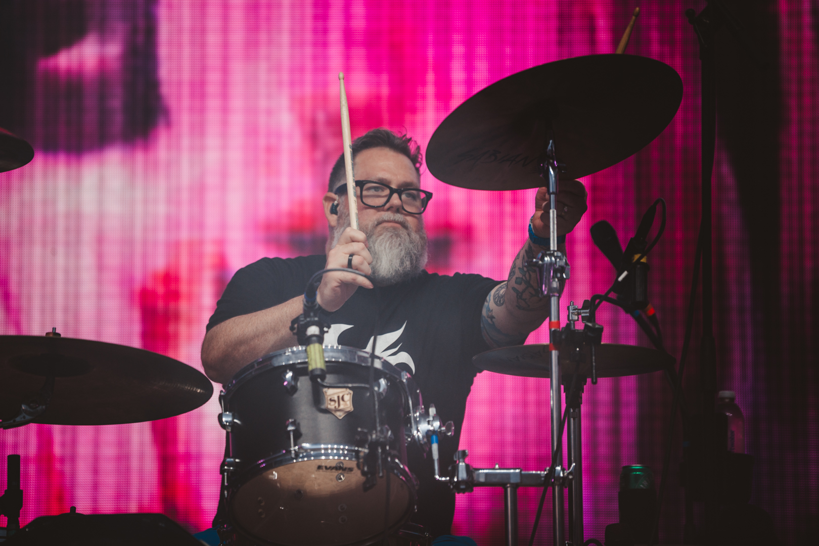 Bearded drummer with glasses performing on stage with pink backdrop