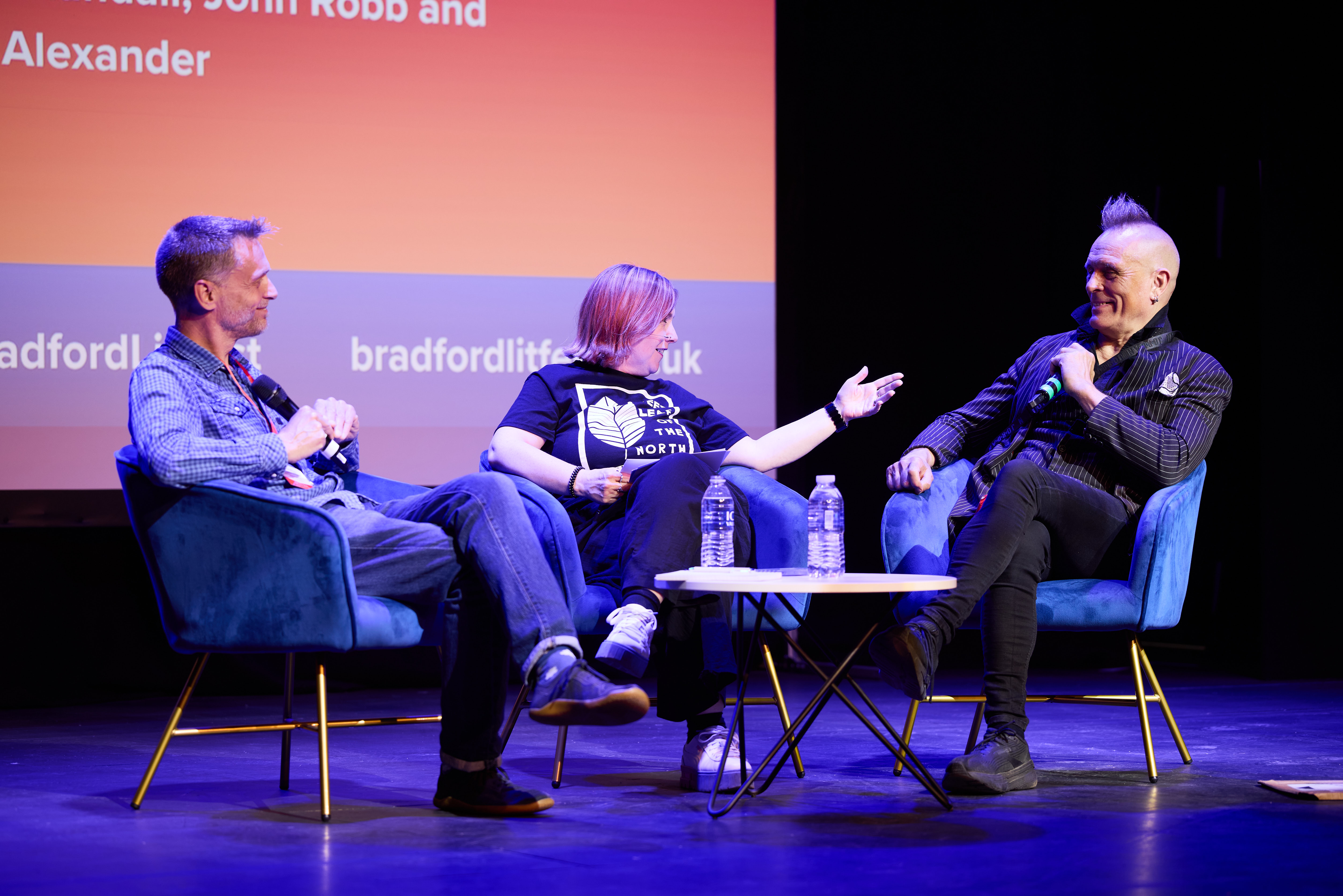 Three people conversing on stage, seated on blue velvet chairs