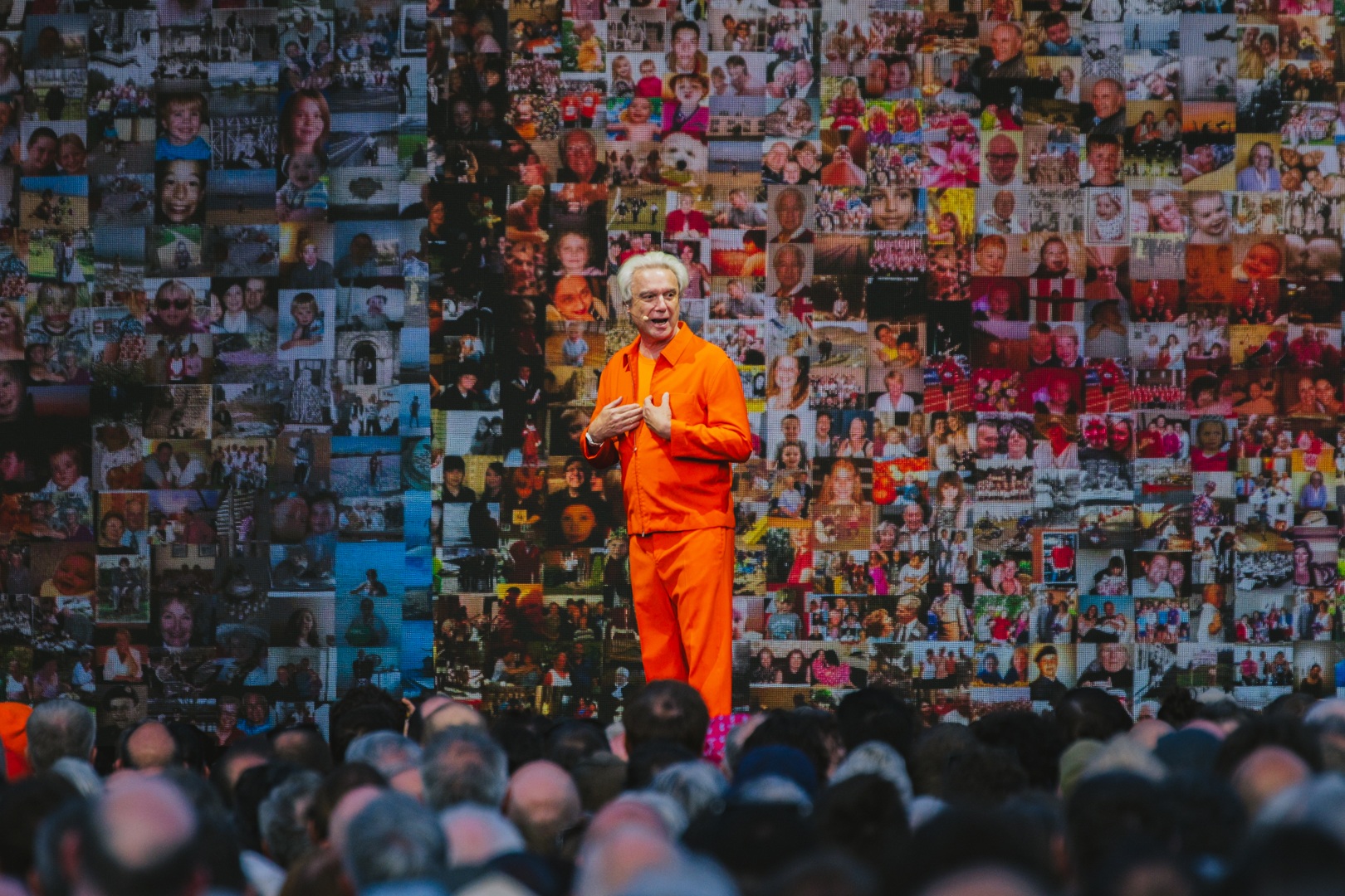 Man in orange jumpsuit speaking on stage with photo collage backdrop