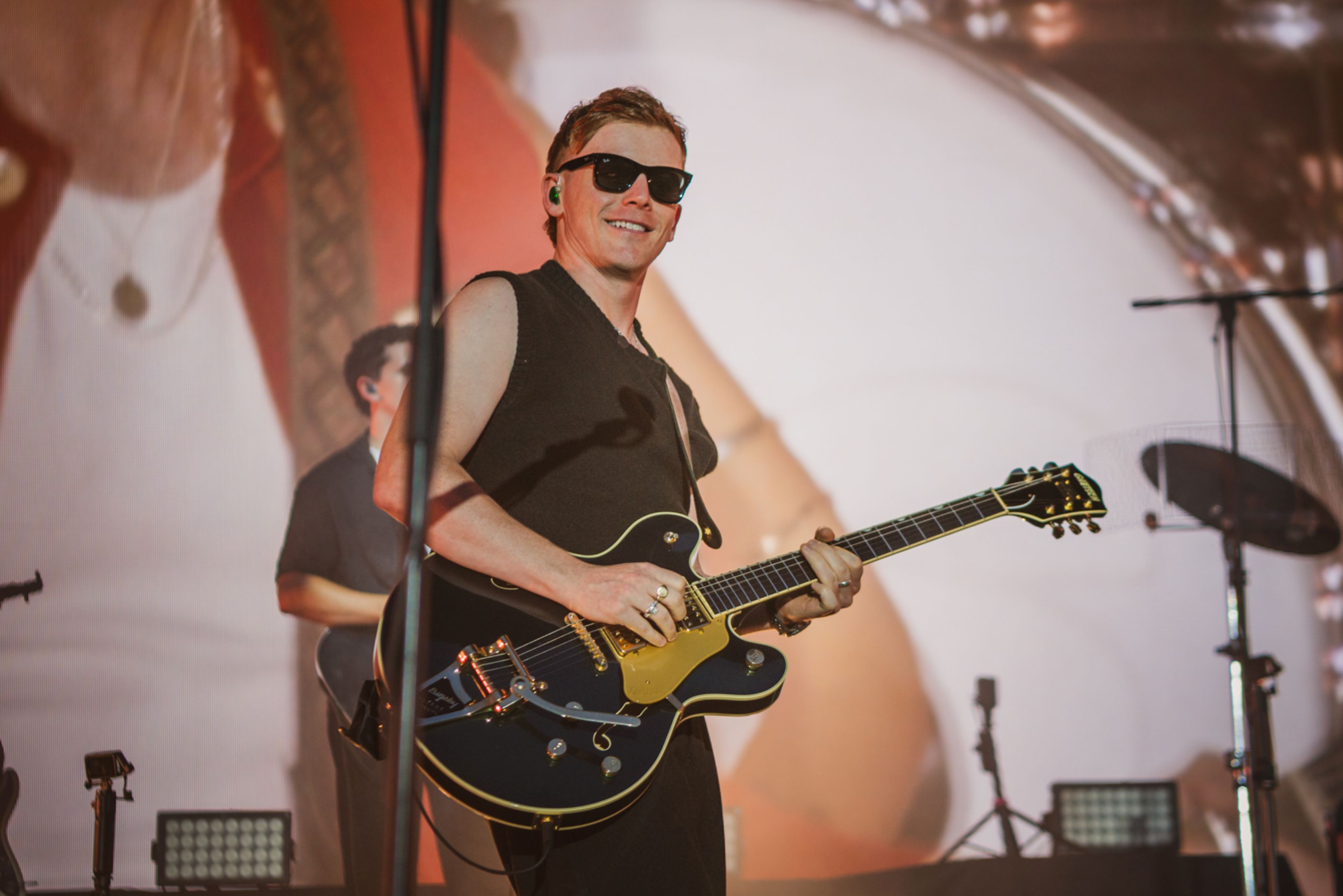 Guitarist in sunglasses performing on stage with a large screen backdrop