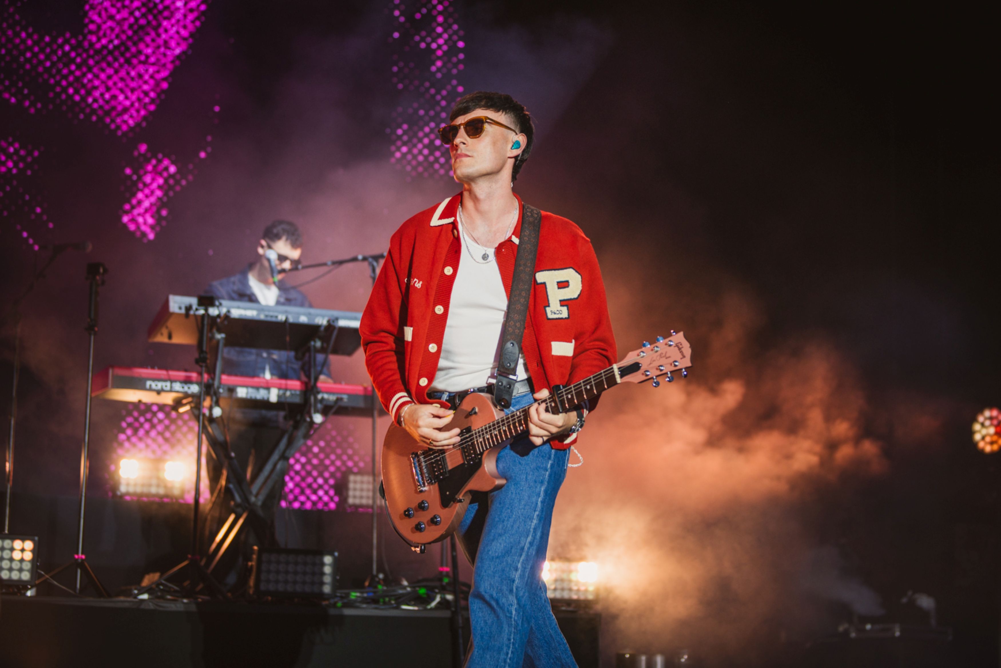 Musician in red jacket playing guitar on stage with keyboardist