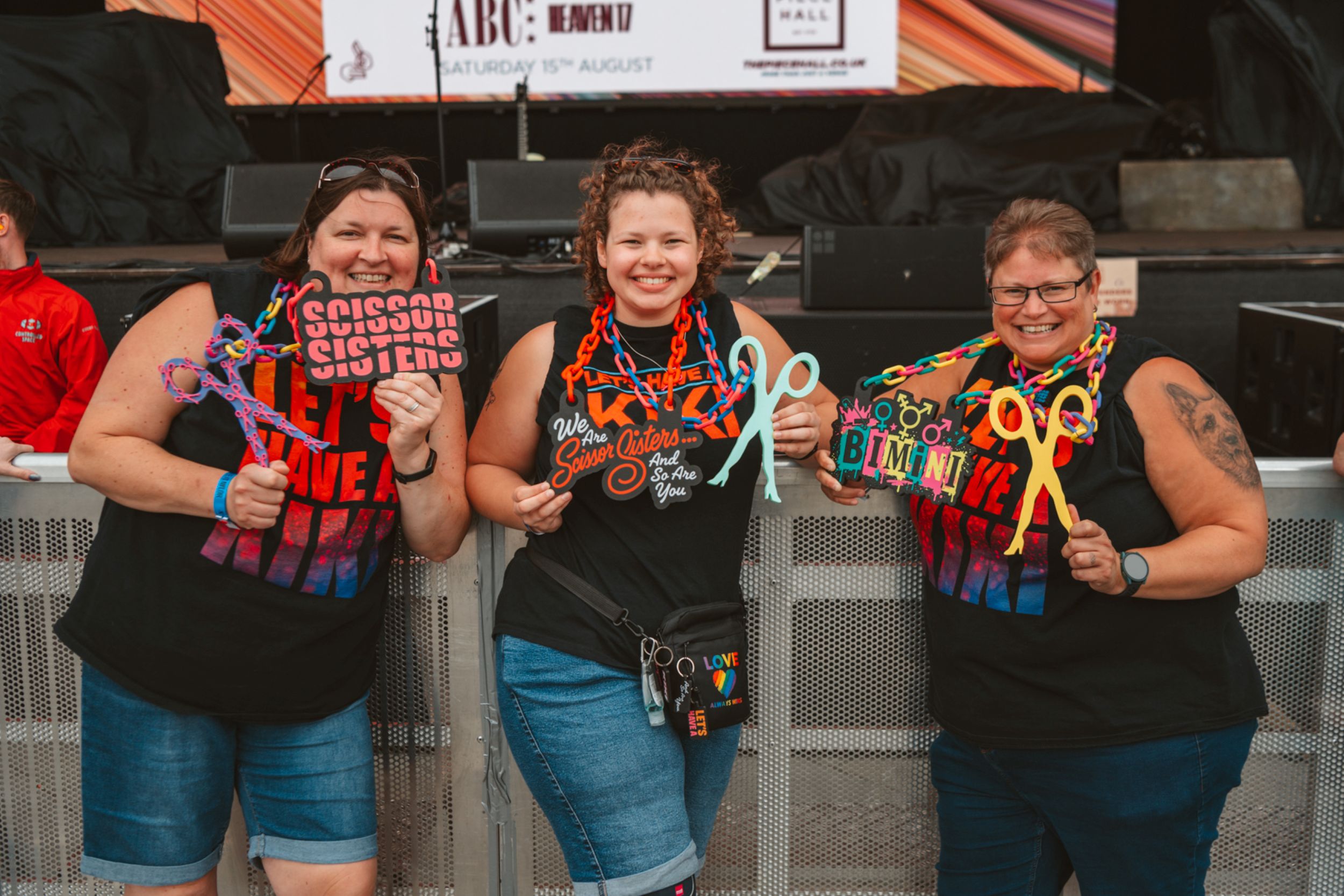 Three smiling women wearing festival shirts holding colourful scissors