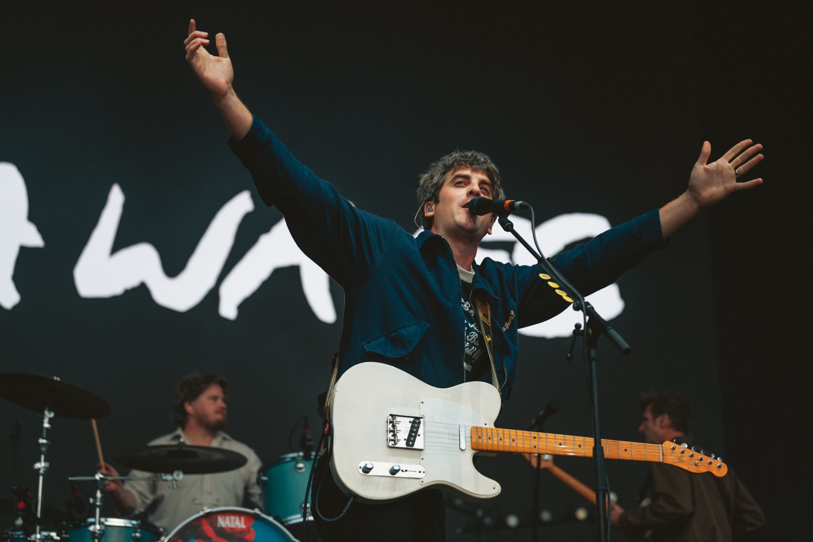 Musician on stage with guitar, arms raised, drums in background