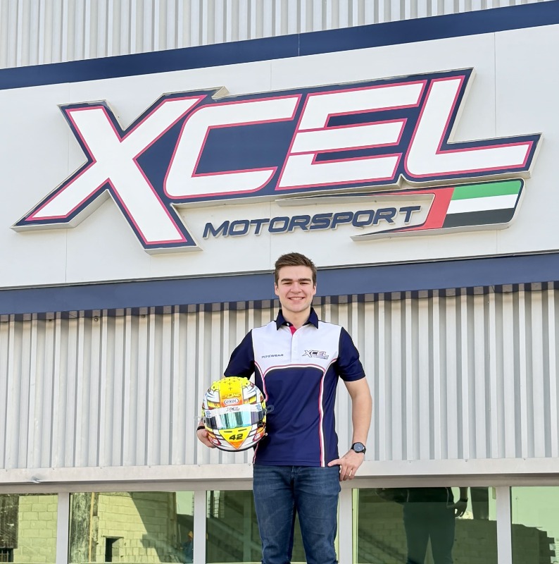 Young man in a racing shirt holds a helmet while standing in front of the XCEL Motorsport sign.