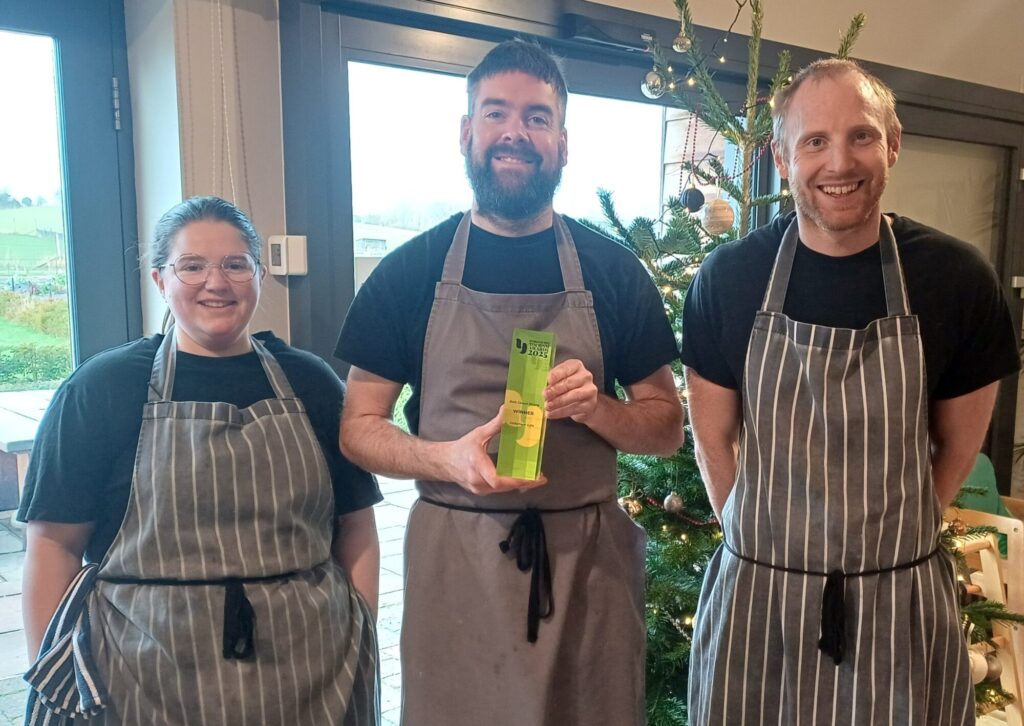 Three smiling staff members in striped aprons stand together, holding an award in front of a festive decorated tree.
