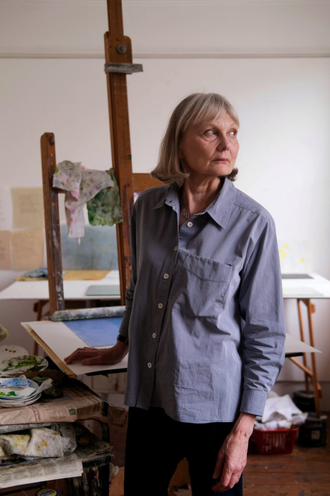 A woman with shoulder-length hair stands thoughtfully in an artist's studio, surrounded by canvases and art materials.