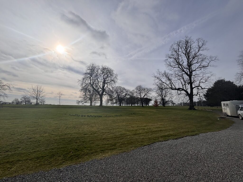Expansive green lawn under a cloudy sky, with scattered trees and a gravel path leading to a white horse trailer.