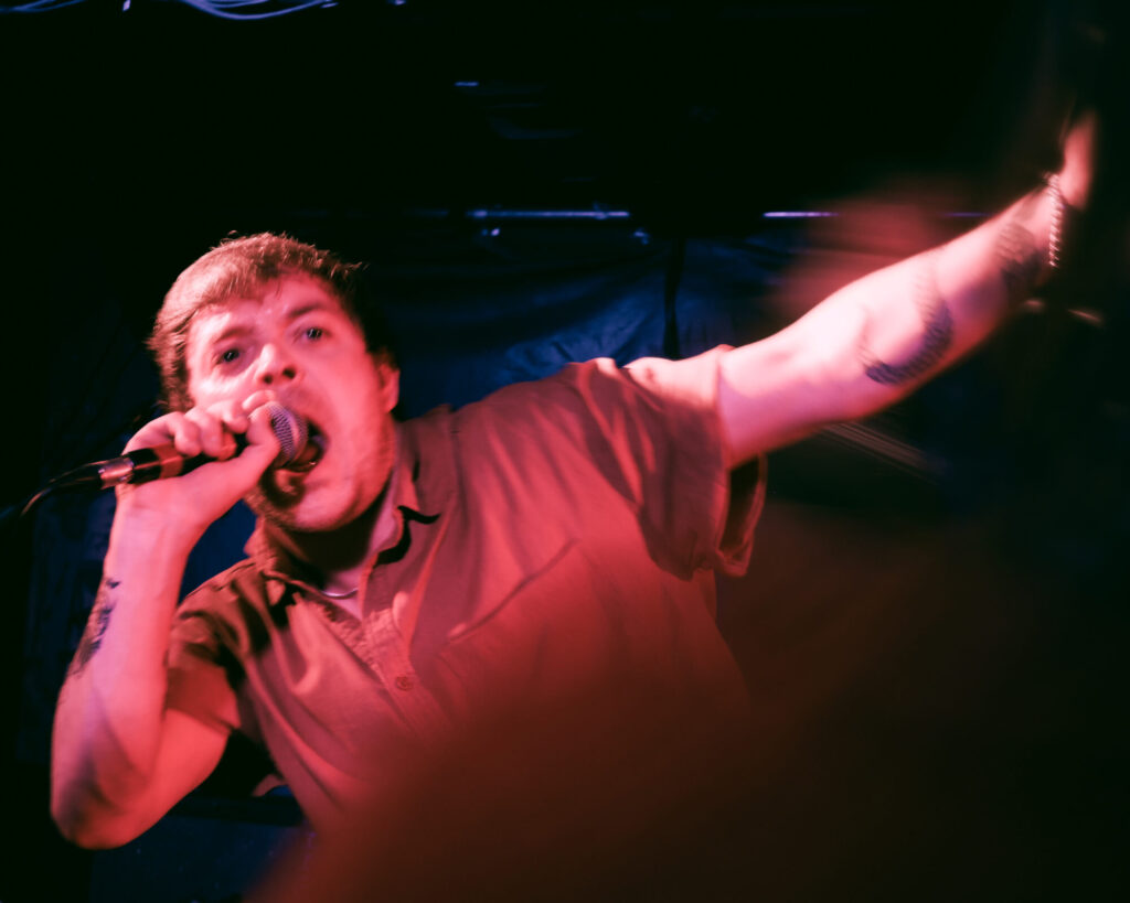 A passionate vocalist performs on stage, gripping a microphone with one arm raised, bathed in vibrant red and blue lights.