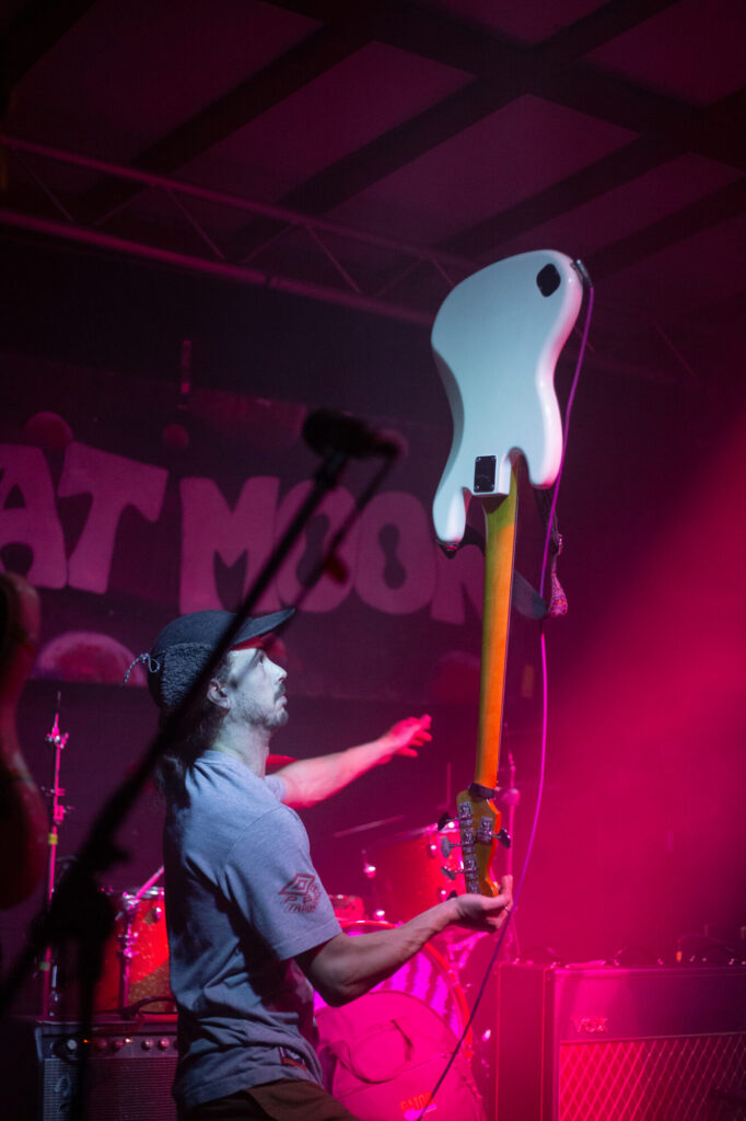 A guitarist joyfully holds a white electric bass guitar aloft, illuminated by vibrant pink stage lights.
