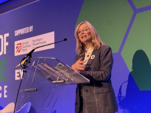 A woman in a grey suit speaks at a podium, with branding for various organizations displayed in the background.