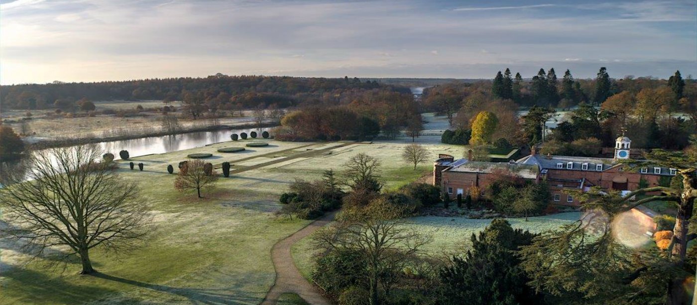 Expansive parkland with frosted grass, a winding path, a lake, and a historic brick building surrounded by trees.