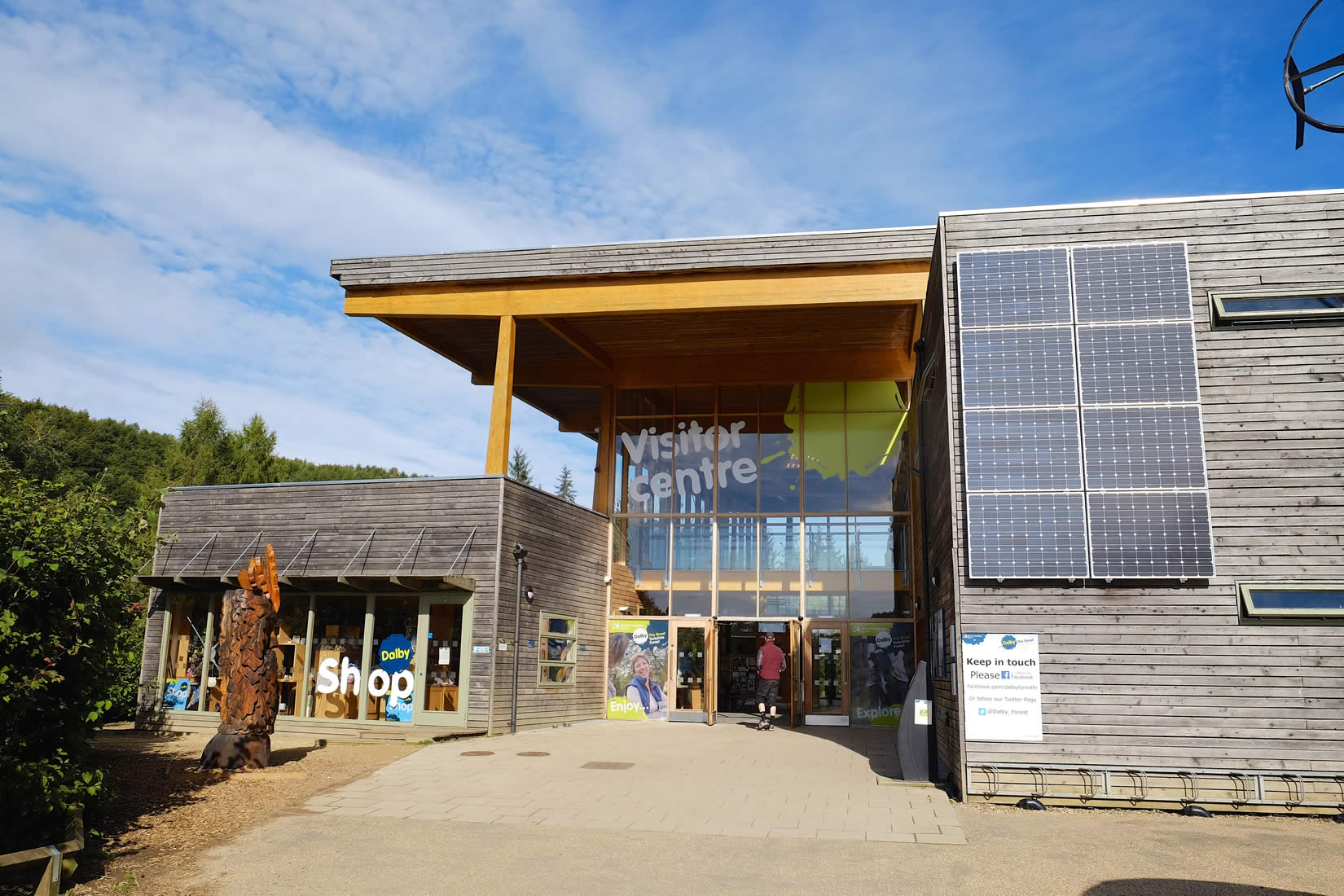 Modern wooden visitor center with large glass windows, solar panels, and people entering under a clear blue sky.