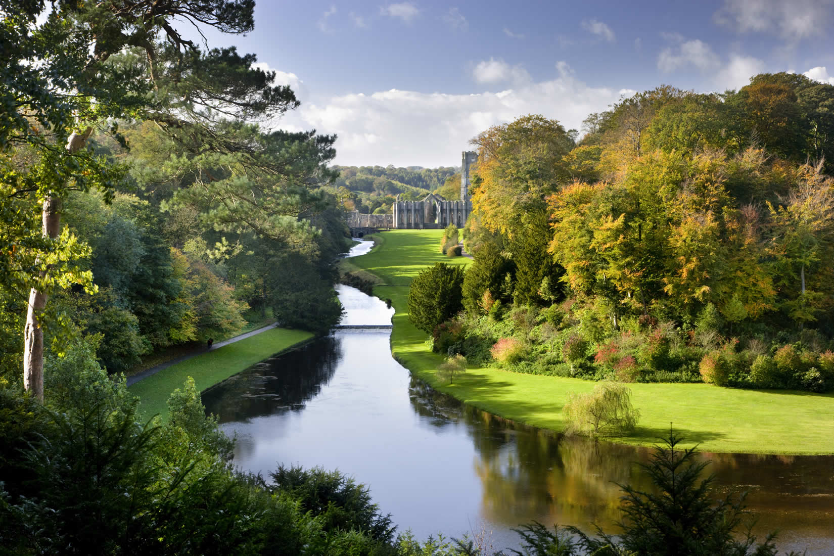 Serene river flanked by lush autumn trees, leading to historic abbey ruins under a clear blue sky.