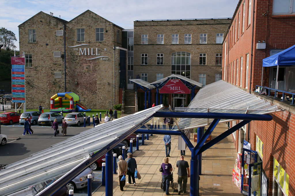 Shoppers walking under a glass canopy towards a large brick factory outlet, with a stone building and a bouncy castle nearby.