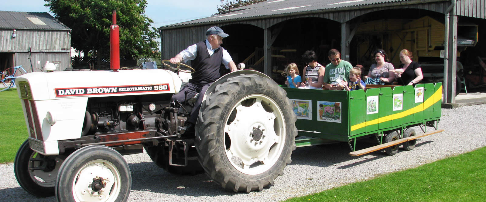 Tractor pulling a green wagon with families and children seated, passing rustic wooden barns on a sunny day.