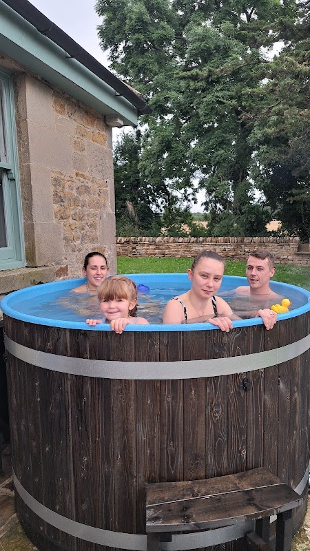 Four people relax in a wooden hot tub outside a stone farmhouse, surrounded by greenery.