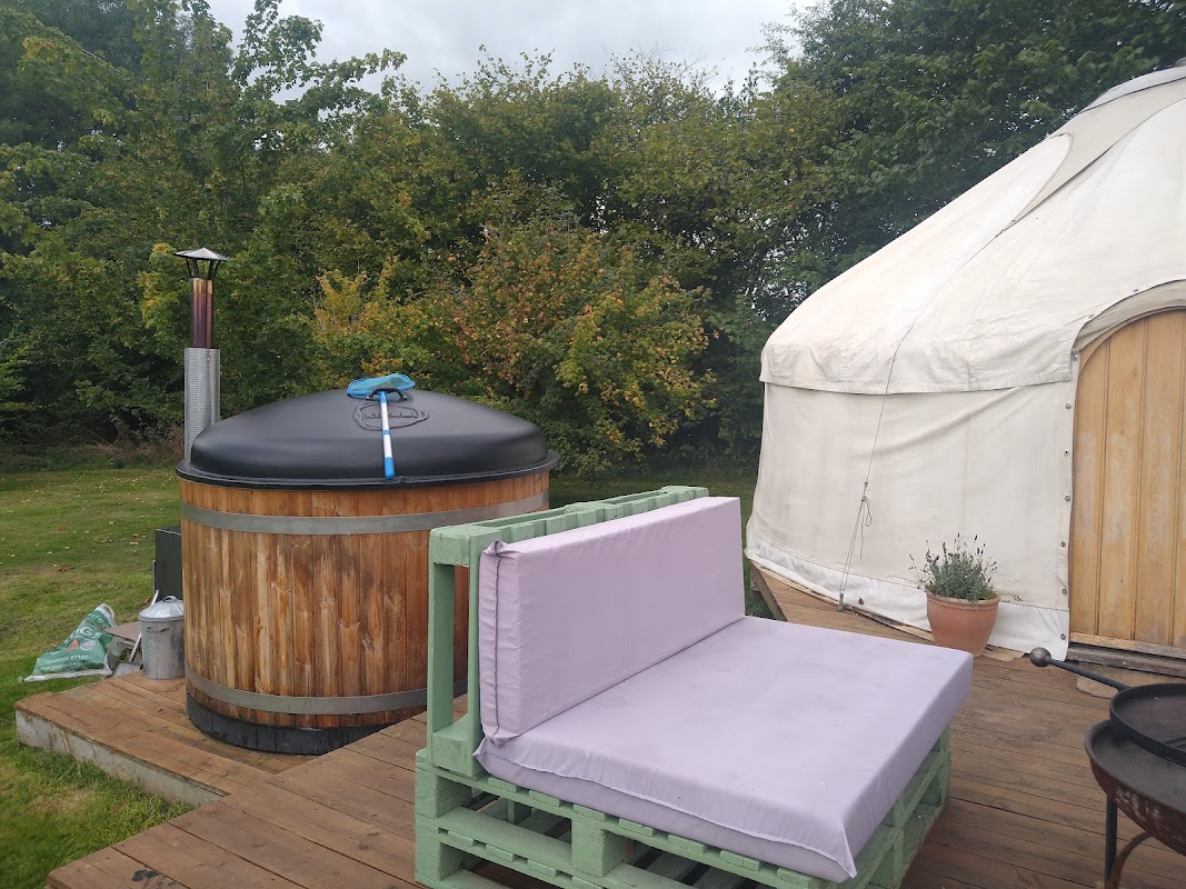 Wooden hot tub with a chimney beside a white yurt and a green bench on a wooden deck, surrounded by trees.
