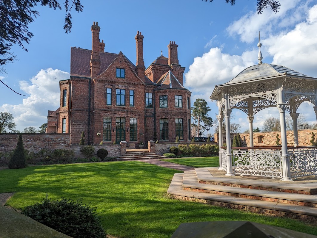 Victorian-style manor with red brick, tall chimneys, and a white gazebo in a landscaped garden under a blue sky.