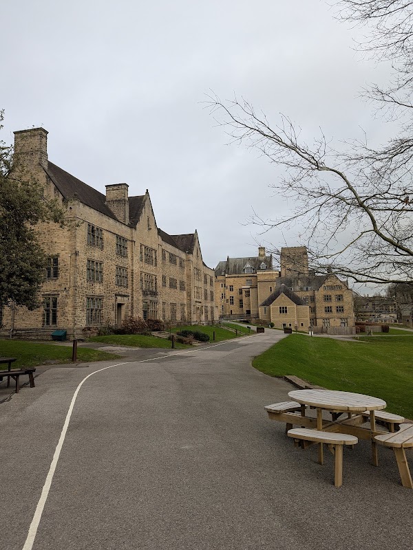 Stone buildings with large windows line a pathway, surrounded by grass and trees, under a cloudy sky.