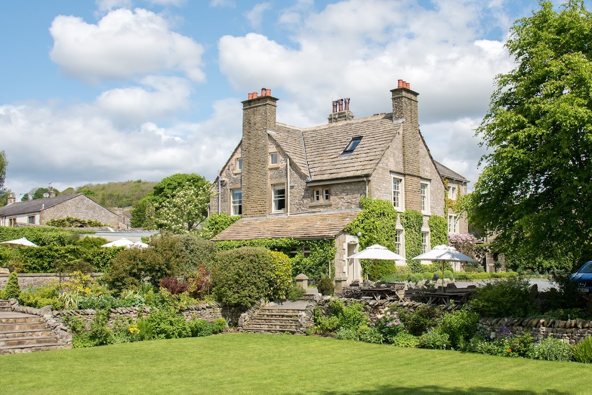 Historic stone venue surrounded by lush gardens and trees, with outdoor seating and a bright blue sky overhead.