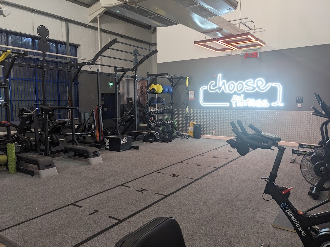 Bright gym interior with workout equipment, weights, and a neon sign reading "choose Fitness."