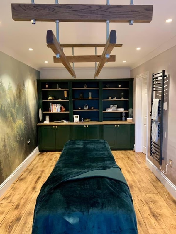 A serene therapy room featuring a massage table, wooden shelving, and calming forest mural on the wall.
