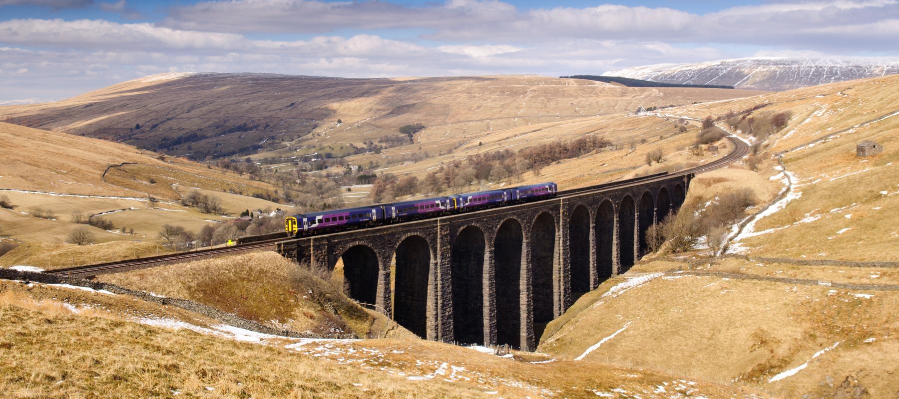 A scenic view of a viaduct with a train crossing, surrounded by rolling hills and fields under a blue sky.